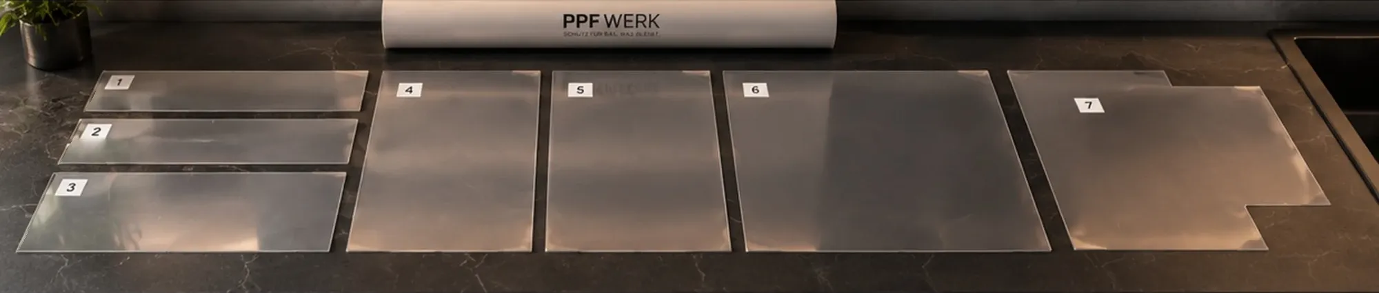 Seven transparent film pieces of different sizes, each with a numbered label, laid side by side on a dark worktop, a film roll behind them.